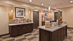 A hotel kitchen with a checkered floor, a central island with kitchen appliances, and various food items on the counter. - Hampton Inn Columbus-South (Grove City)