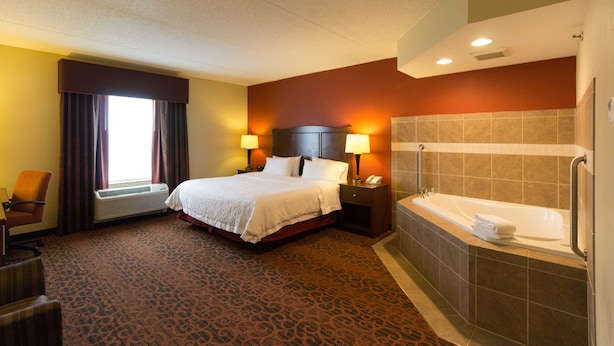 A hotel room with a bed, two bedside lamps, a desk, an air conditioner, a window with curtains, and a bathroom with a bathtub and towel rack. - Hampton Inn Columbus-South (Grove City)