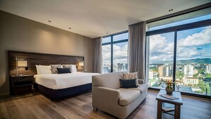 A modern hotel room with a large bed, a sofa, a small table, and a view of the cityscape through large windows.