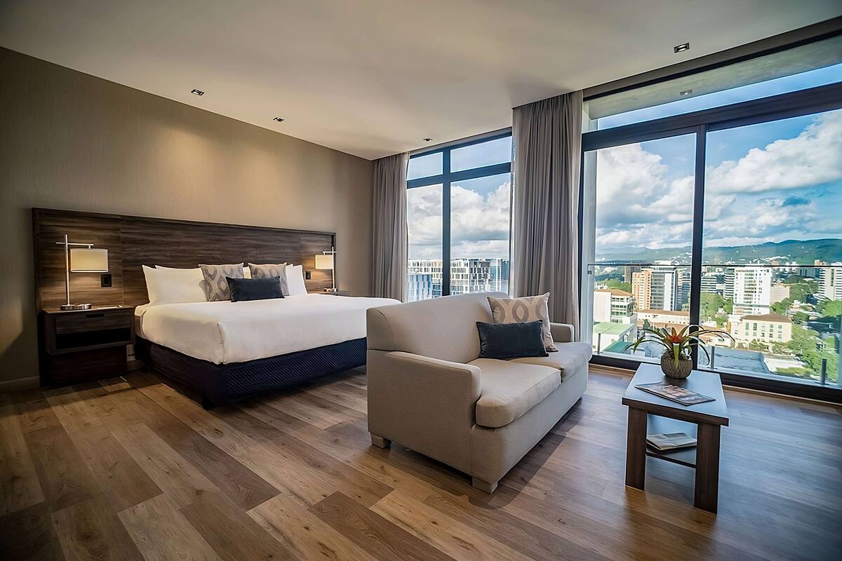 Ein modernes Hotelzimmer mit einem großen Bett, einer Couch, einem kleinen Tisch und Blick auf die Stadt durch große Fenster.