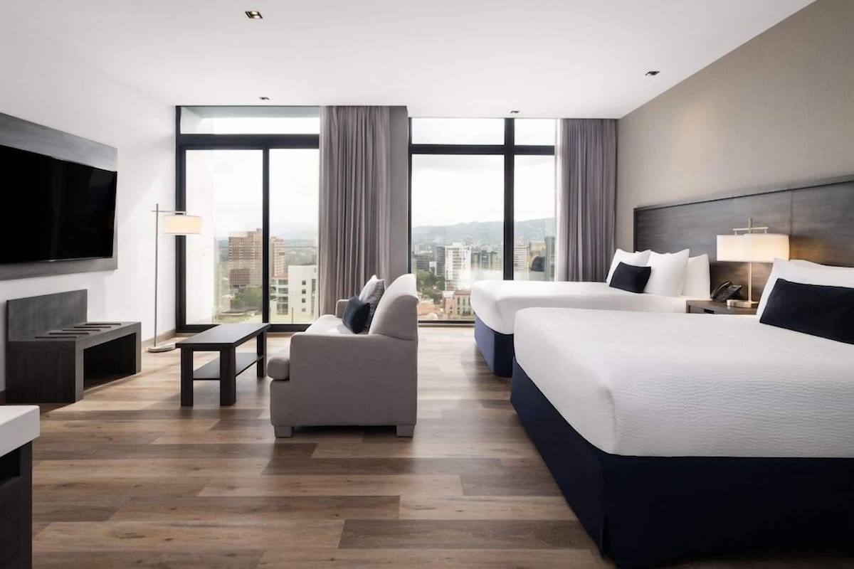 A modern hotel room with a large bed, a sofa, a TV, and a view of the city.