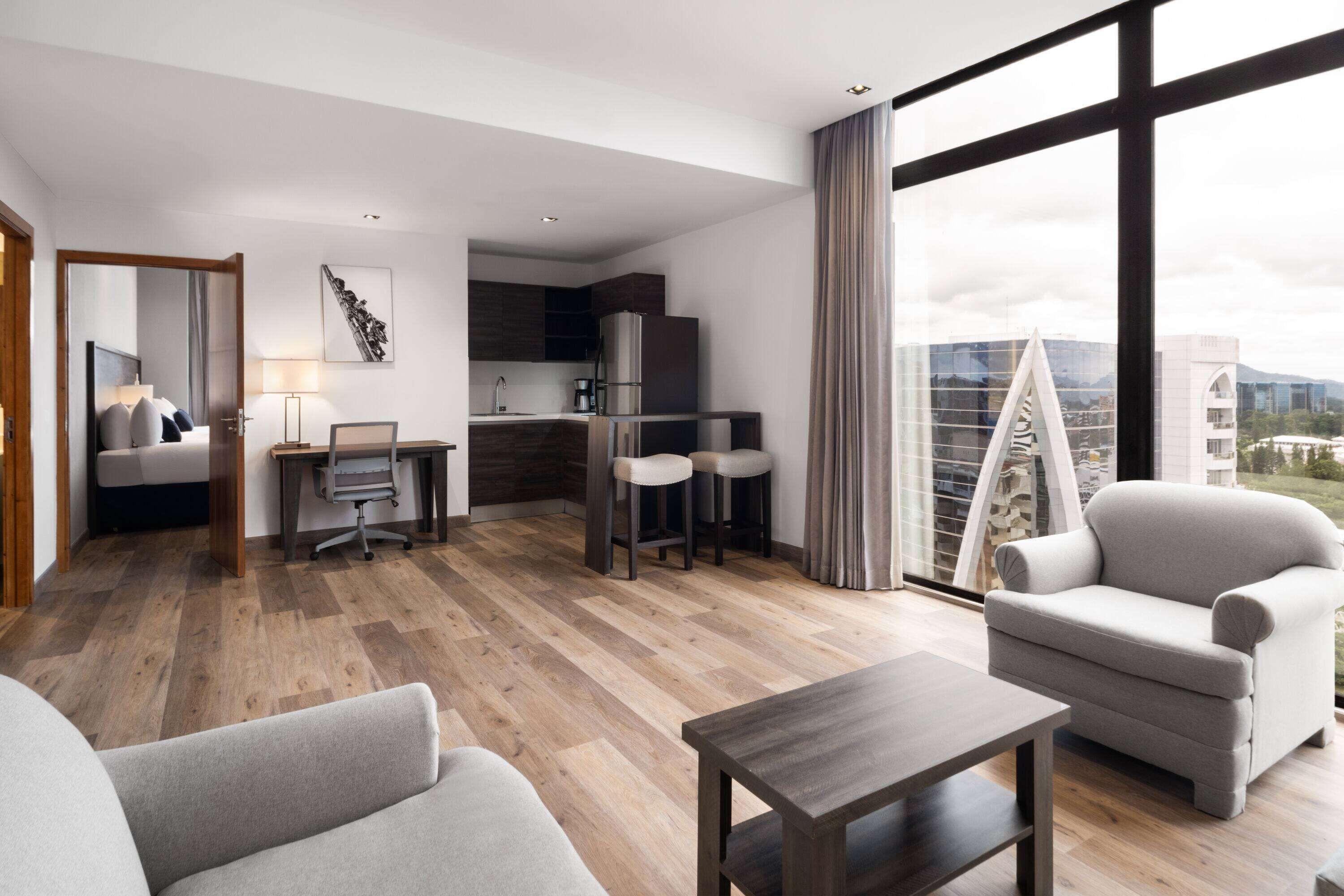 A modern hotel room with a living area, a dining area, and a kitchen. There is a large window with a view of the city.