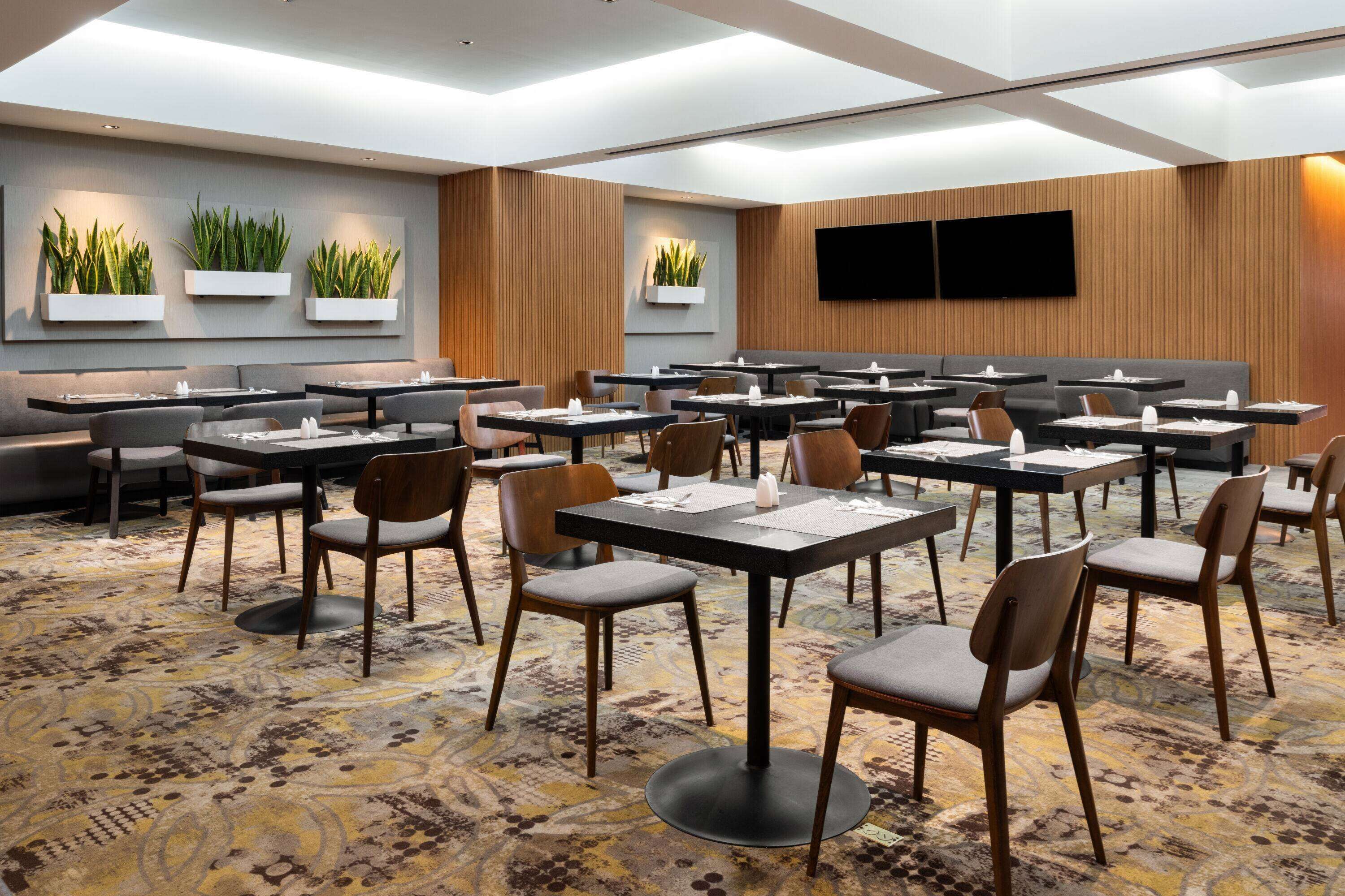 A hotel conference room with multiple tables and chairs, a patterned carpet, and wall-mounted planters with green plants.