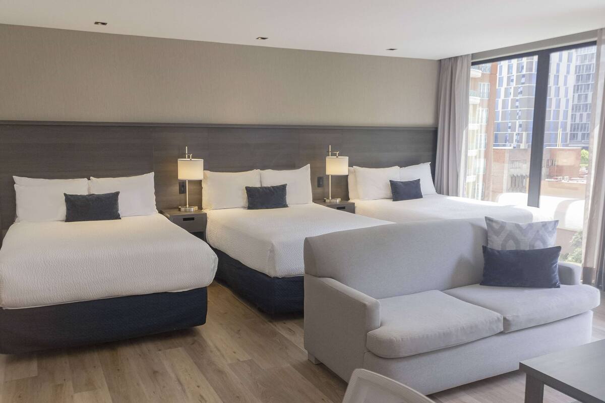 Ein Hotelzimmer mit zwei Betten, einem Schlafsofa und einem großen Fenster mit Blick auf die Stadt.