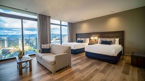 A modern hotel room with two beds, a sofa, a small table, and large windows offering a city view.
