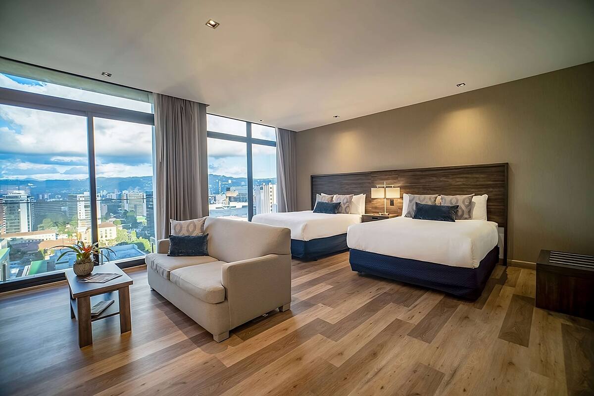 Ein modernes Hotelzimmer mit zwei Betten, einer Couch, einem kleinen Tisch und großen Fenstern mit Blick auf die Stadt.