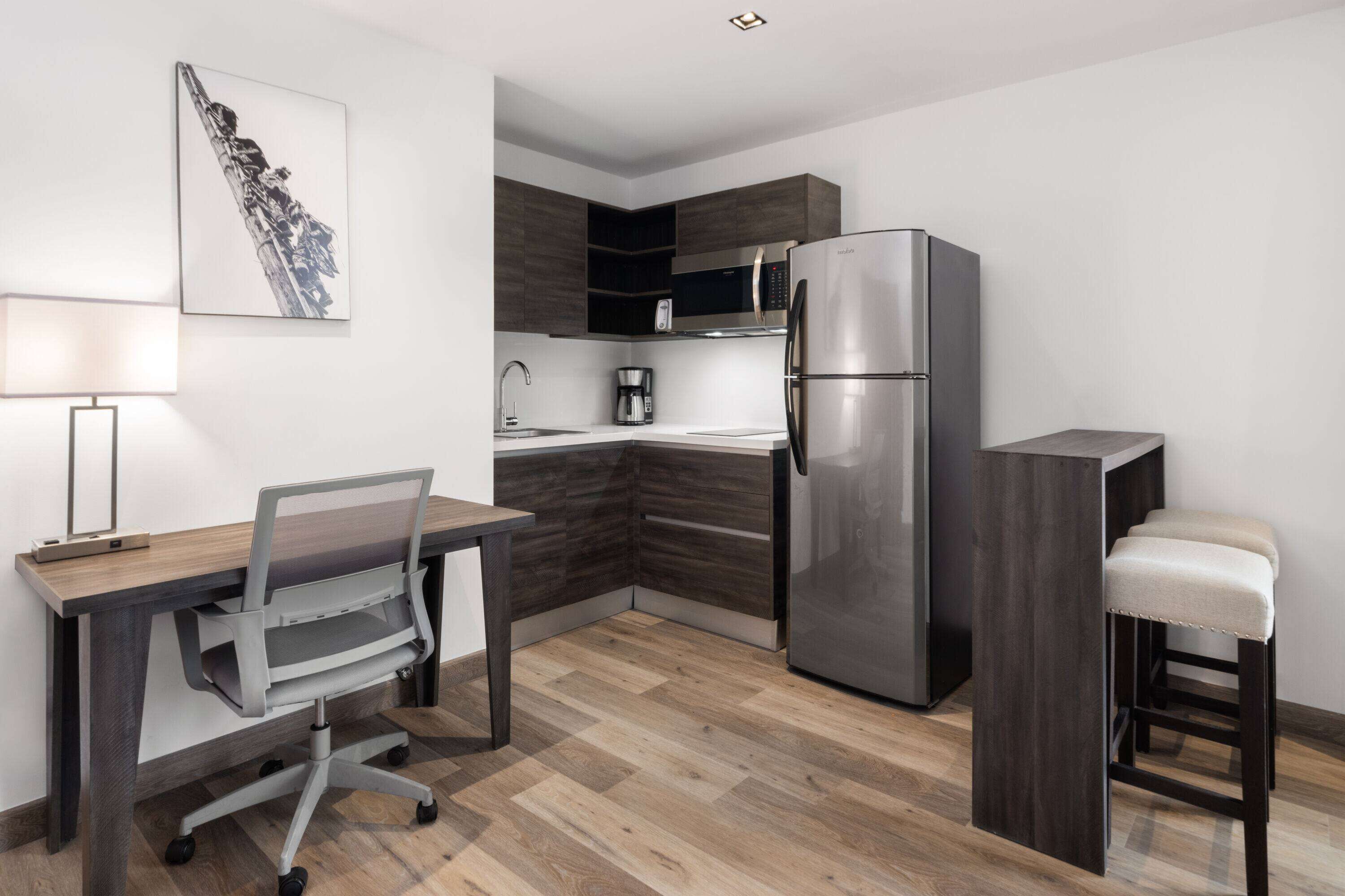 Una cocina moderna con microondas integrado, refrigerador y cafetera. Hay un escritorio de madera con una silla y un taburete.