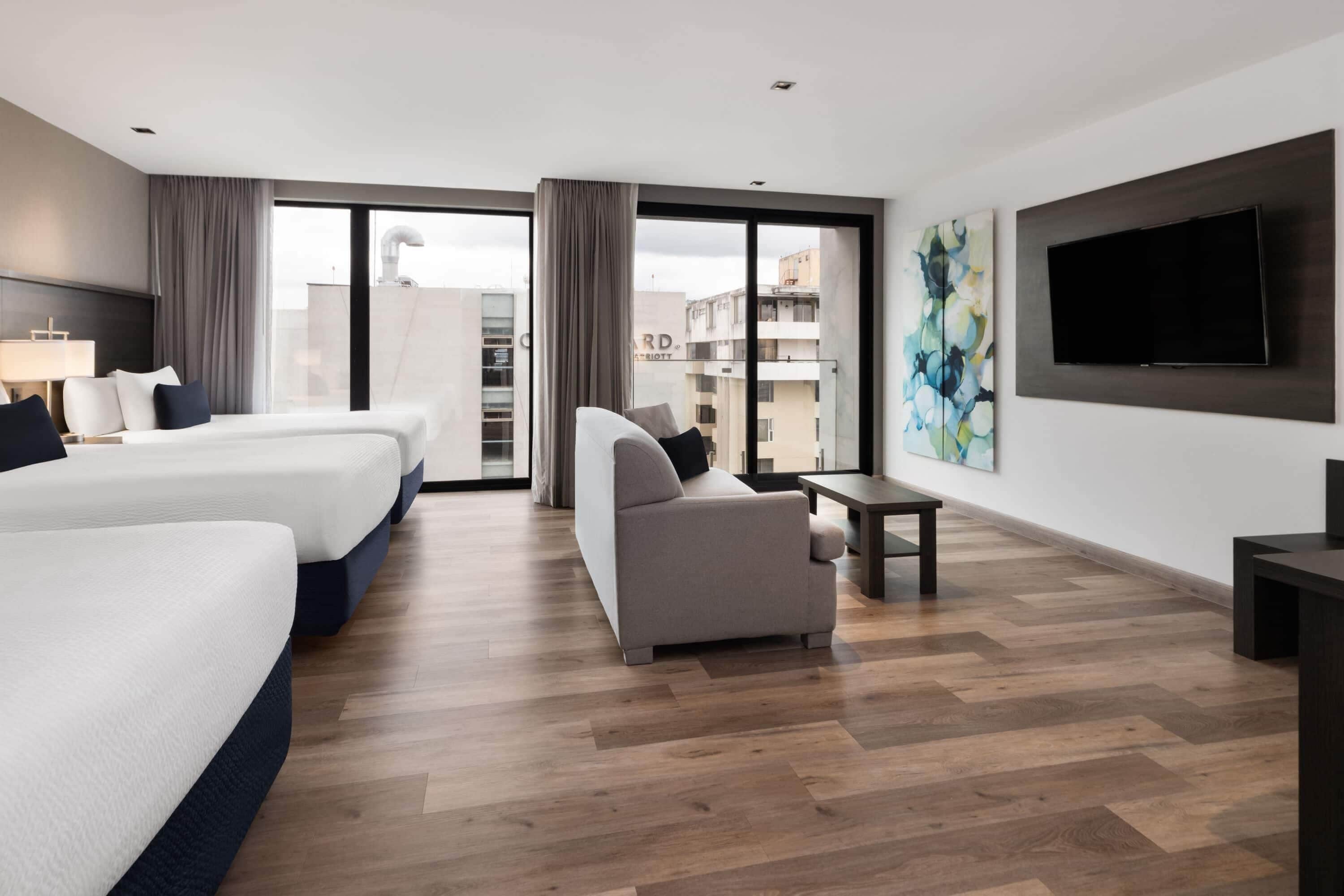 A hotel room with two beds, a sofa, a TV, and a large window with a view of buildings.