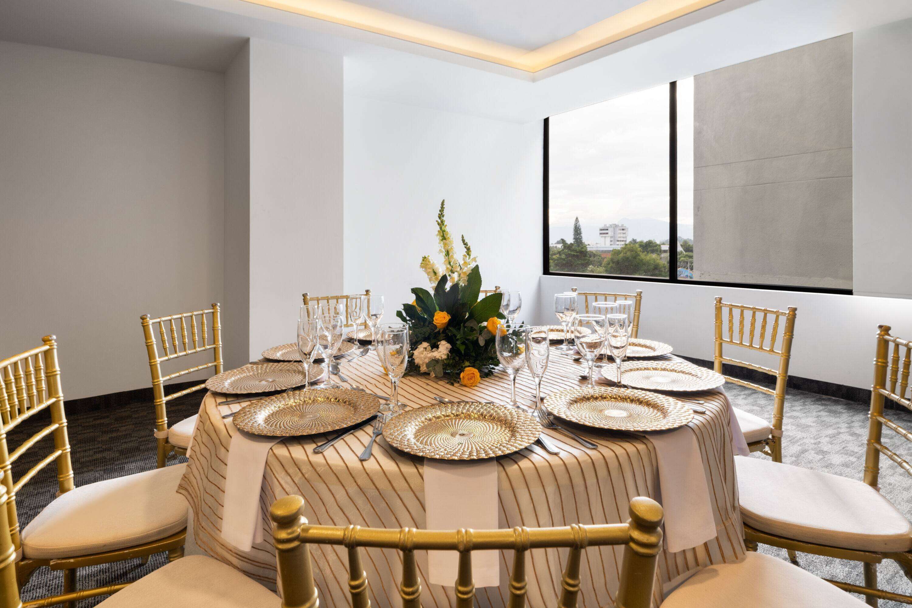 Comedor con una mesa redonda dispuesta para seis personas, con sillas doradas, manteles blancos y un centro de mesa floral.