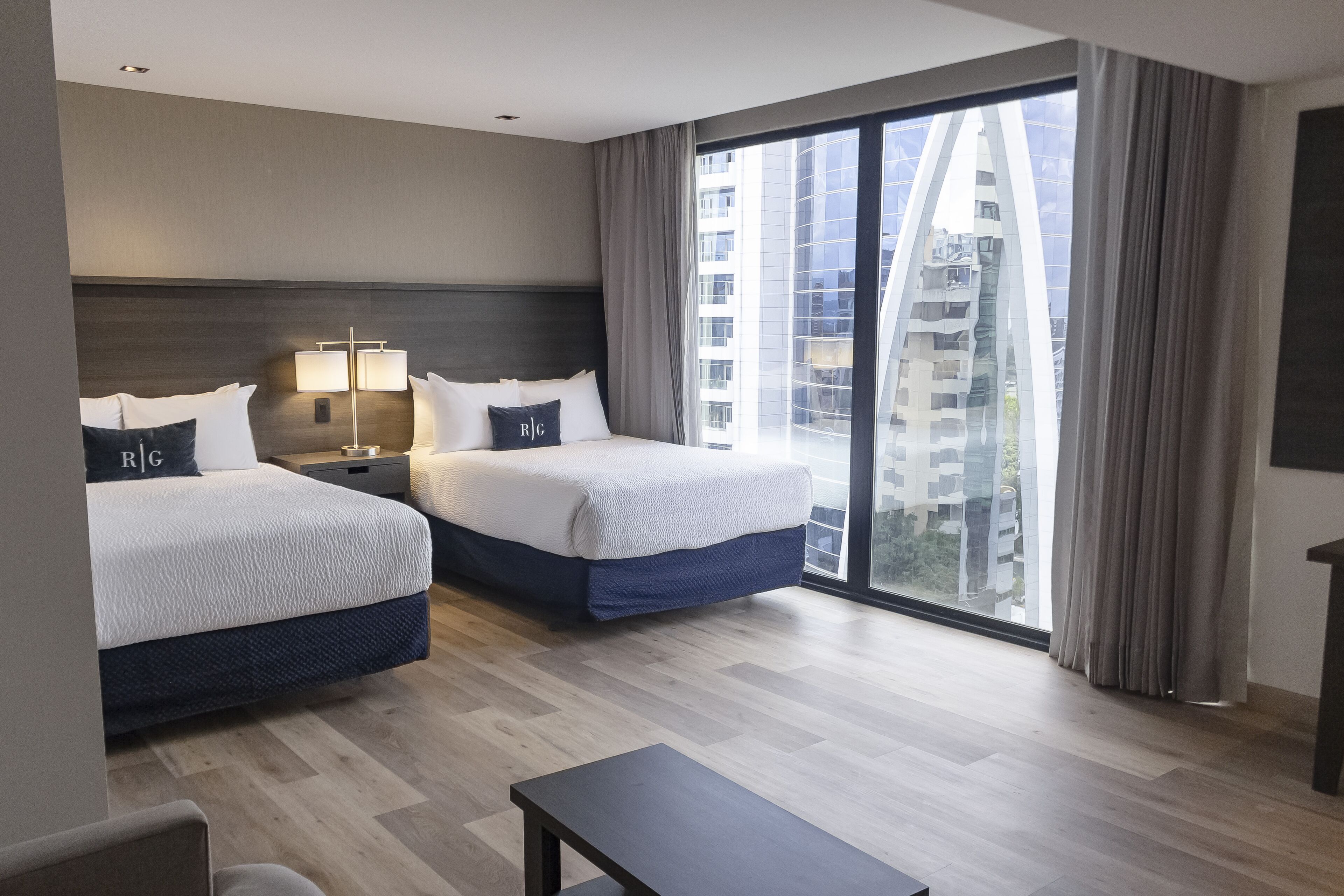 A hotel room with two beds, a nightstand, a lamp, a desk, and a large window with curtains.
