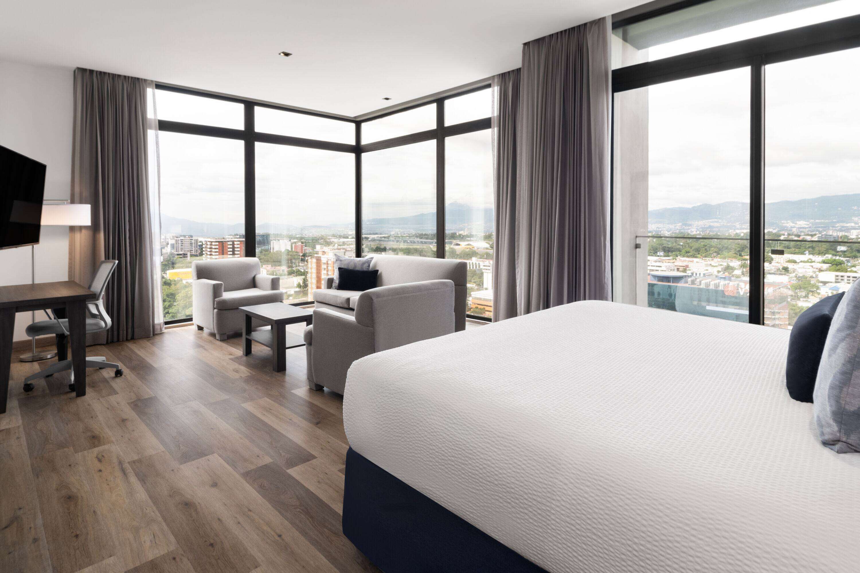 A modern hotel room with a large bed, a desk, a sofa, and a view of the city.
