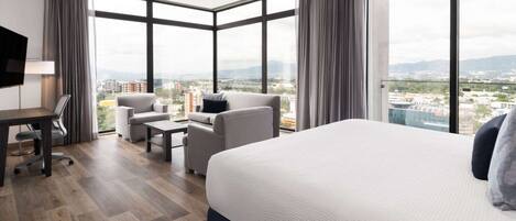 A modern hotel room with a large bed, a desk, a sofa, and a view of the city.