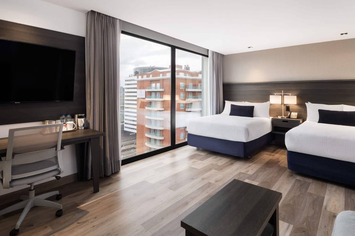 Ein Hotelzimmer mit einem großen Bett, einem Schreibtisch mit Stuhl, einem Fernseher und Blick auf ein Gebäude durch ein großes Fenster.
