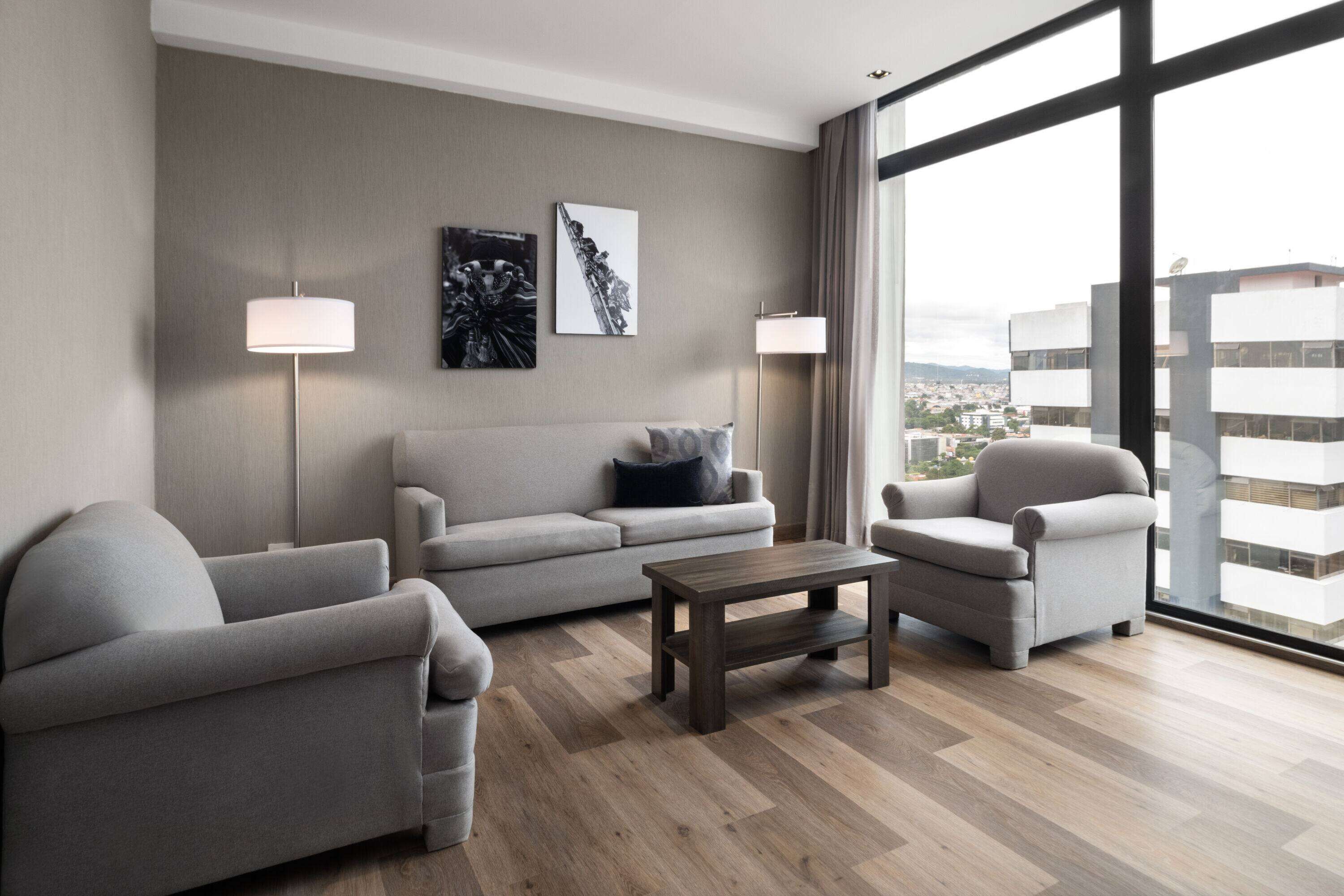 A modern living room with a sofa, two armchairs, a coffee table, and floor lamps. There are two wall-mounted artworks and a large window providing a view of buildings.