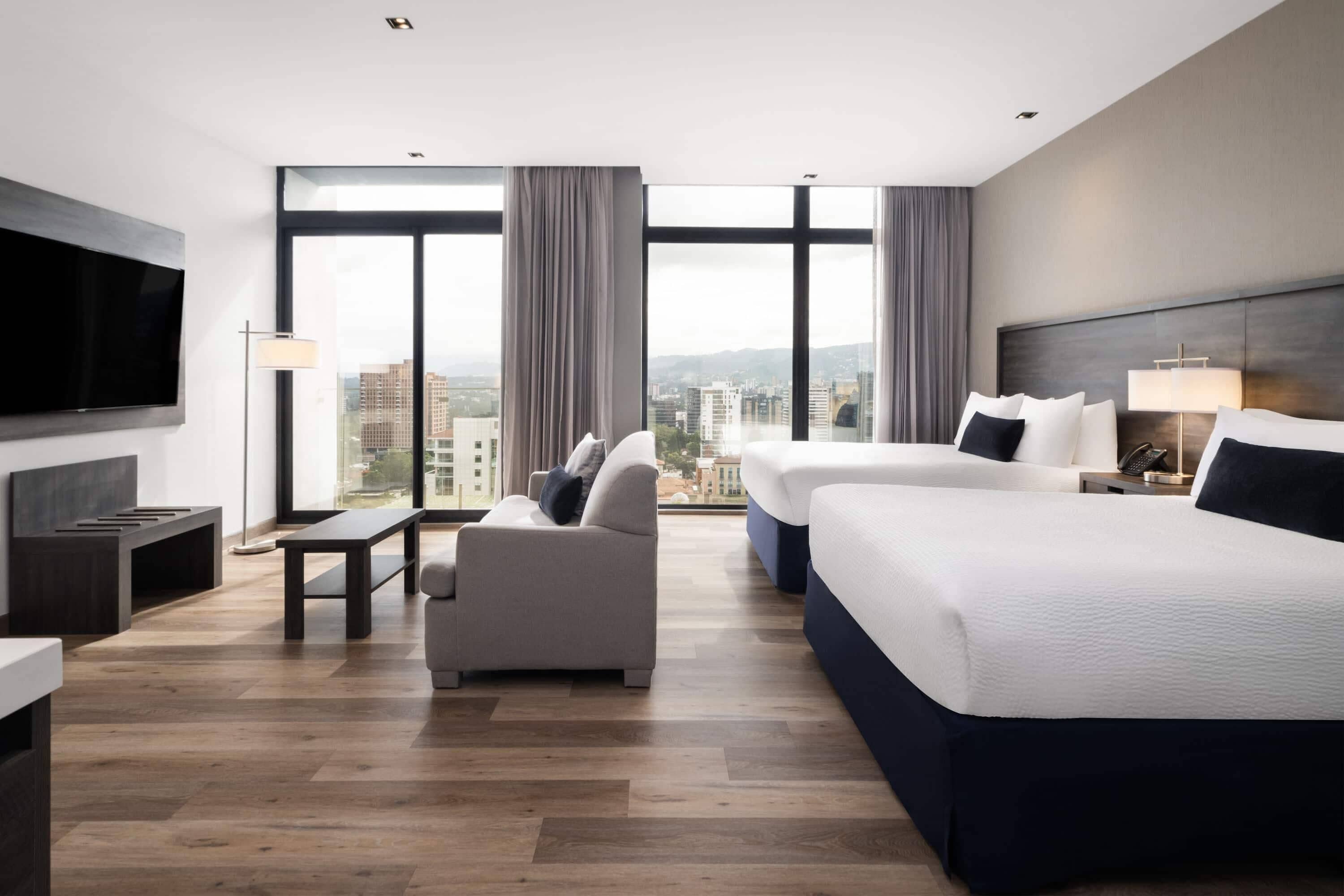 A modern hotel room with a large bed, a sofa, a TV, and a city view.