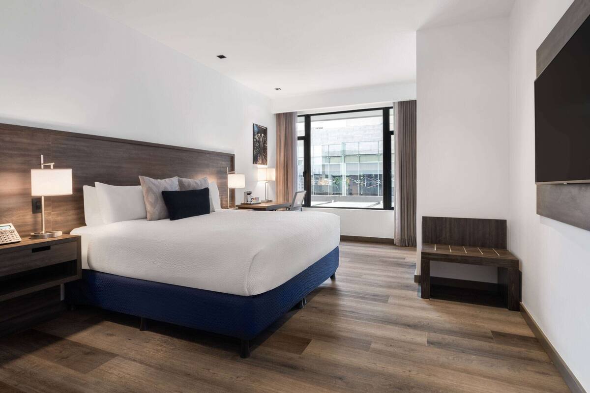 Ein modernes Hotelzimmer mit einem großen Bett, Nachttischlampen, einem Nachttisch, einer kleinen Bank und einem großen Fenster mit Blick.