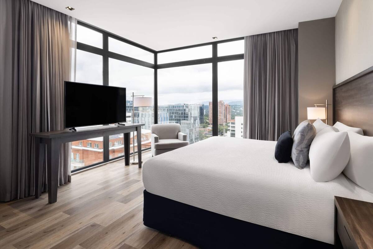 Ein modernes Hotelzimmer mit einem großen Bett, einem Flachbildfernseher, einem Schreibtisch und einem Sessel. Das Zimmer verfügt über große Fenster mit Blick auf die Stadt.