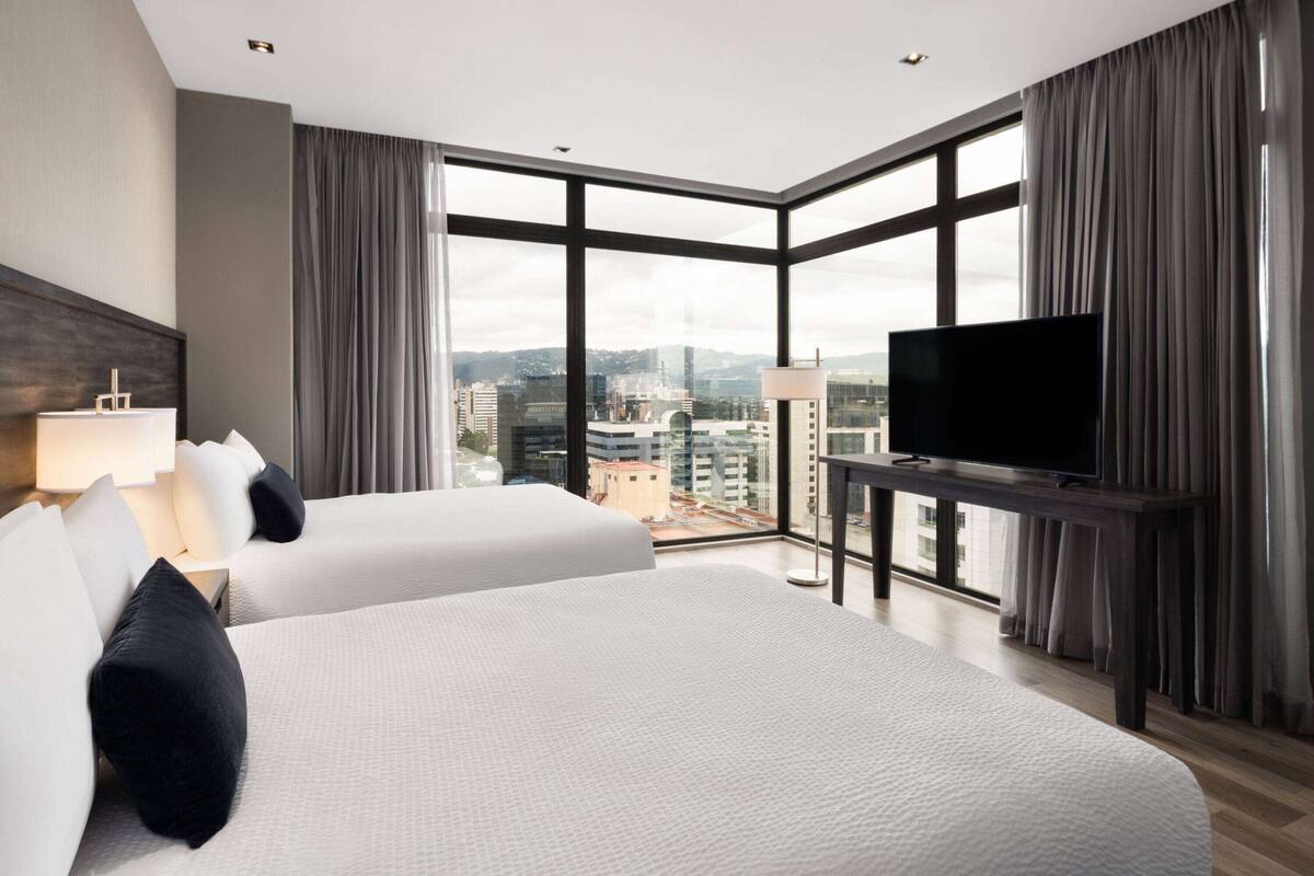 Ein Hotelzimmer mit zwei Betten, einem Fernseher und großen Fenstern mit Blick auf die Stadt.
