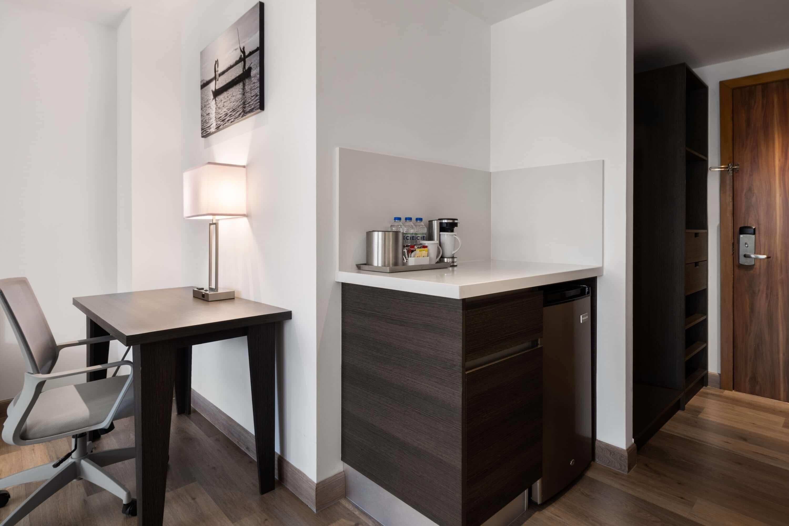 A hotel room with a desk, chair, lamp, coffee maker, and a small kitchenette.