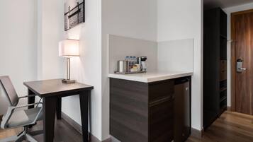 A hotel room with a desk, chair, lamp, coffee maker, and a small kitchenette.