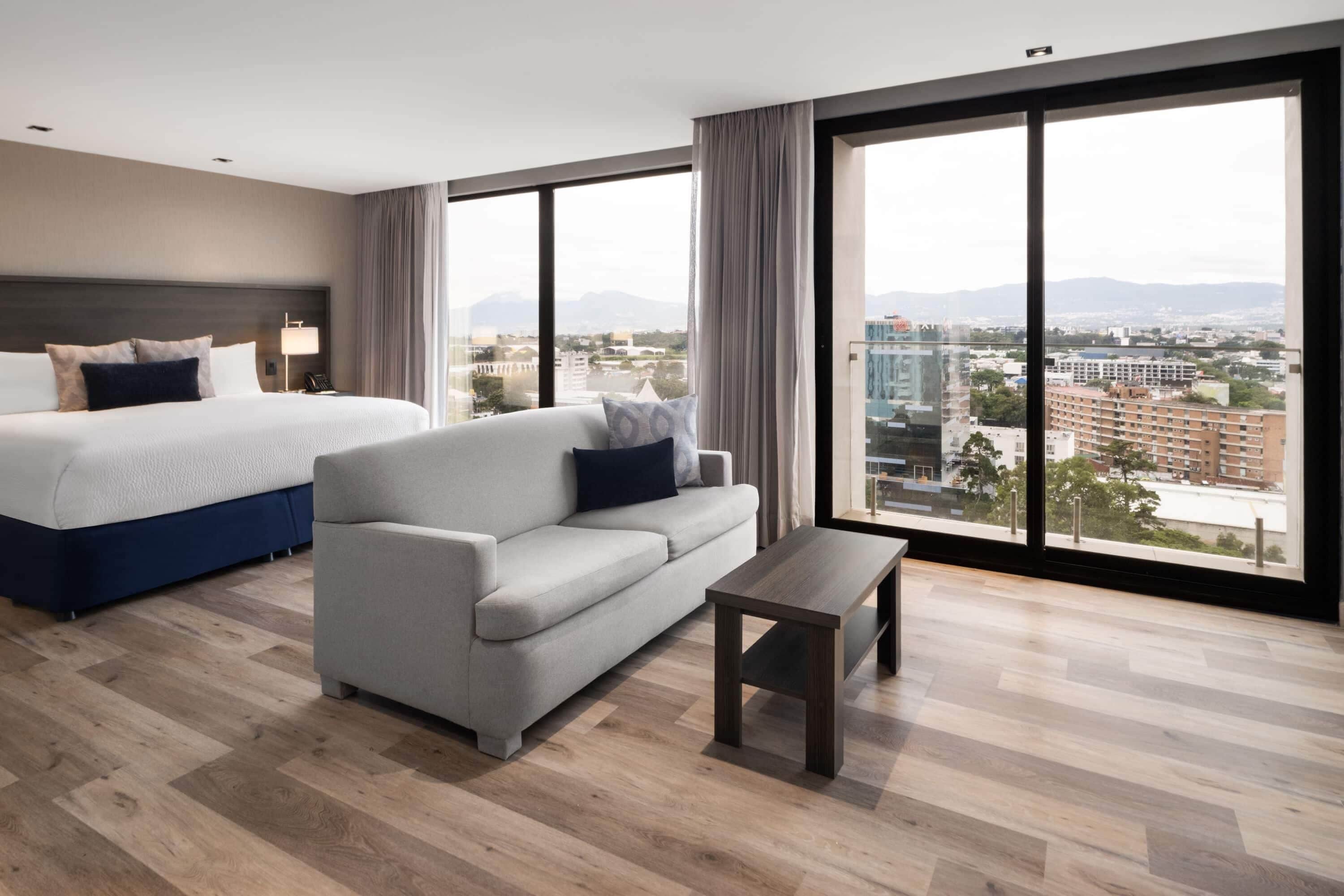 A modern hotel room with a large bed, a sofa, and a small table. The room has large windows offering a city view.