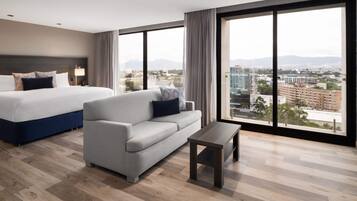 A modern hotel room with a large bed, a sofa, and a small table. The room has large windows offering a city view.