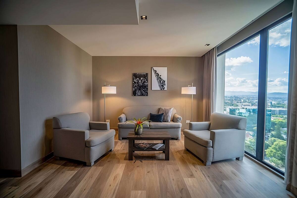 Ein modernes Wohnzimmer mit einer Sofagarnitur, zwei Sesseln, einem Couchtisch und zwei Stehlampen. An der Wand befinden sich zwei gerahmte Kunstwerke und ein großes Fenster mit Blick auf die Stadt.