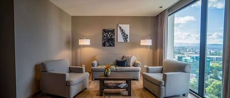 A modern living room with a sofa set, two armchairs, a coffee table, and two floor lamps. There are two framed artworks on the wall and a large window offering a city view.