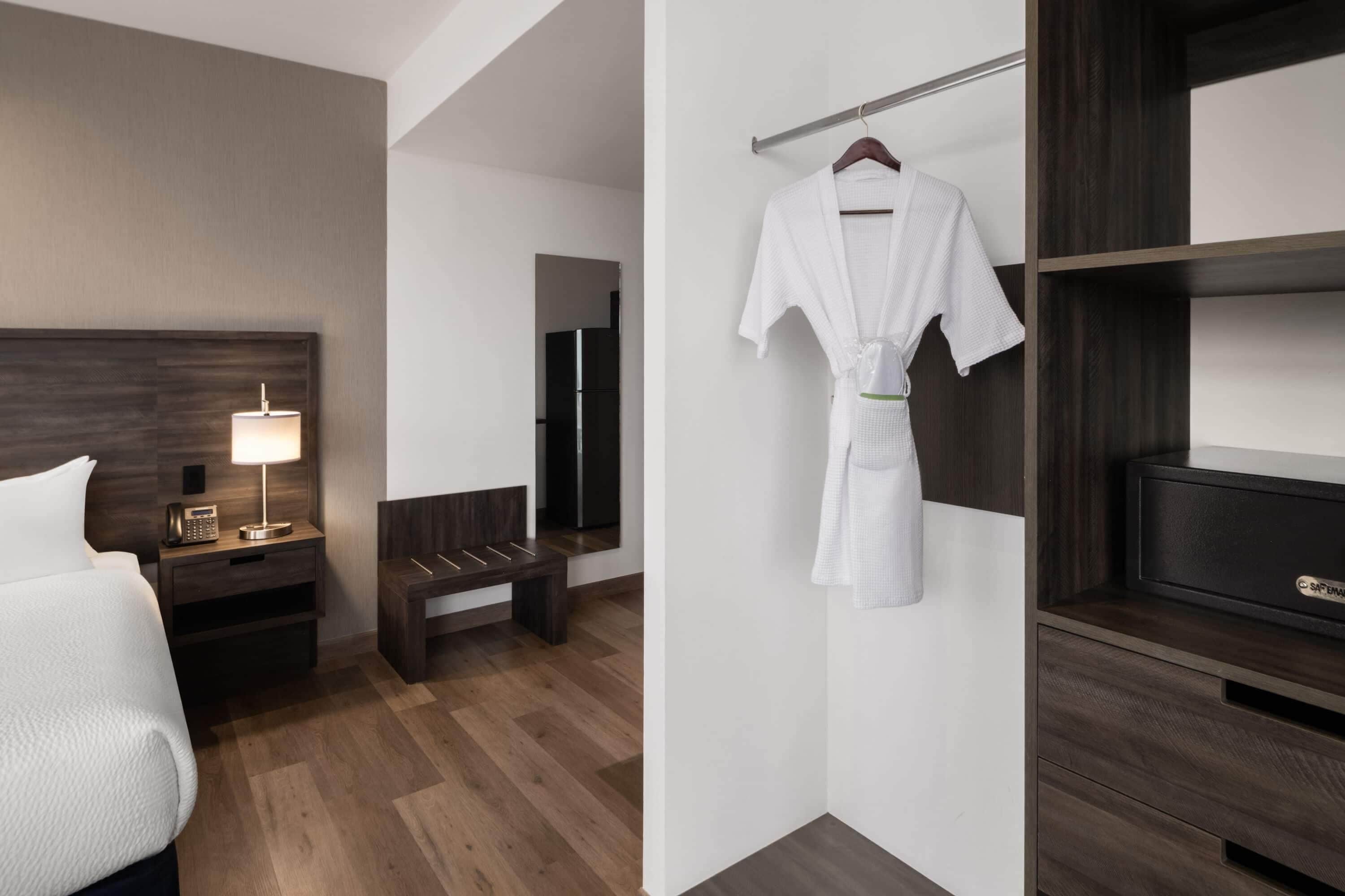 A hotel room with a bed, bedside table with a lamp, a bench, a robe hanging on a rack, and a TV on a wooden shelf.