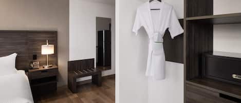 Habitación de hotel con cama, mesita de noche con lámpara, un banco, bata colgada en un perchero y un televisor sobre una repisa de madera.