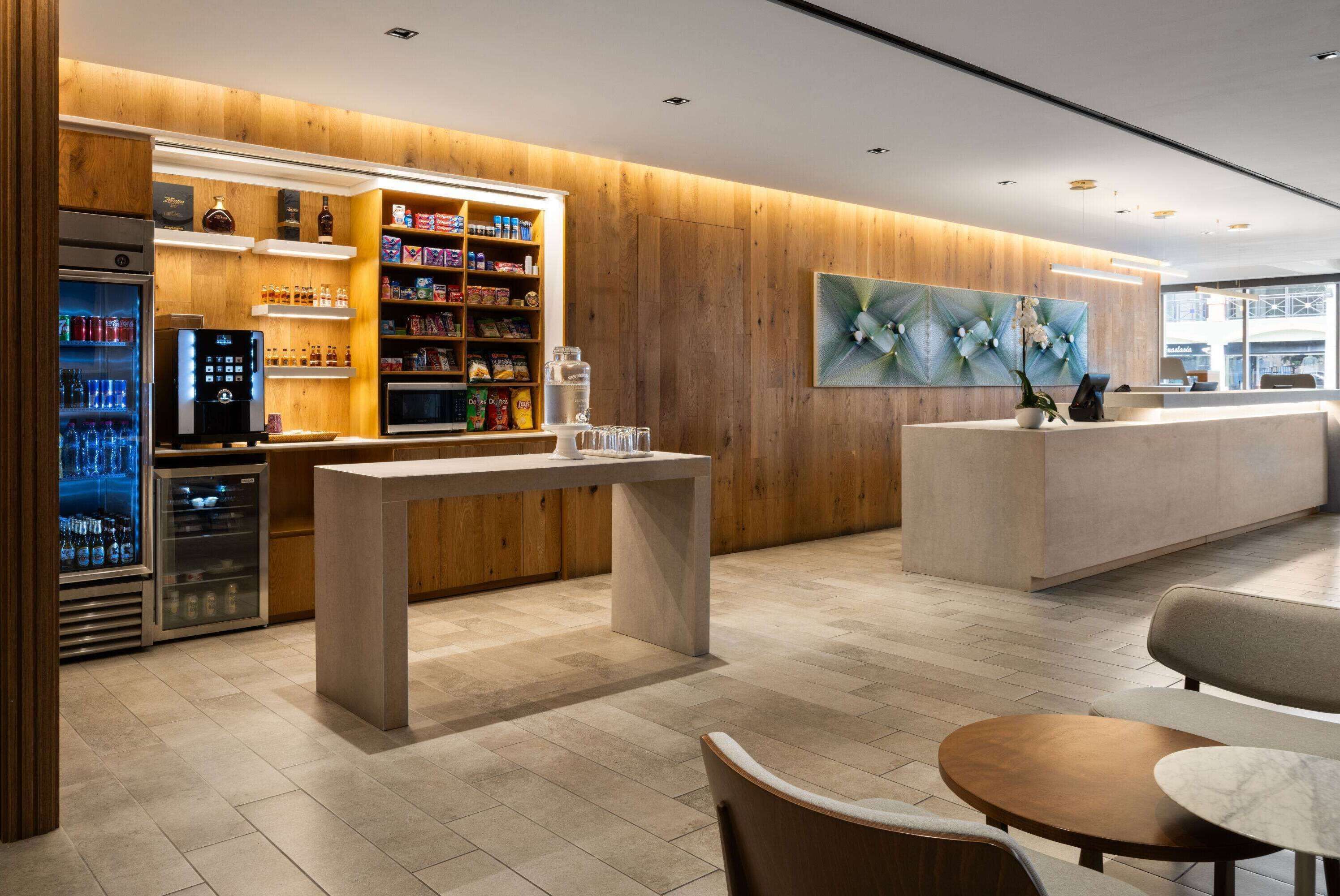 Un moderno vestíbulo de hotel con un mostrador de recepción de madera, un dispensador de bebidas refrigeradas y una zona de estar con mesas redondas.