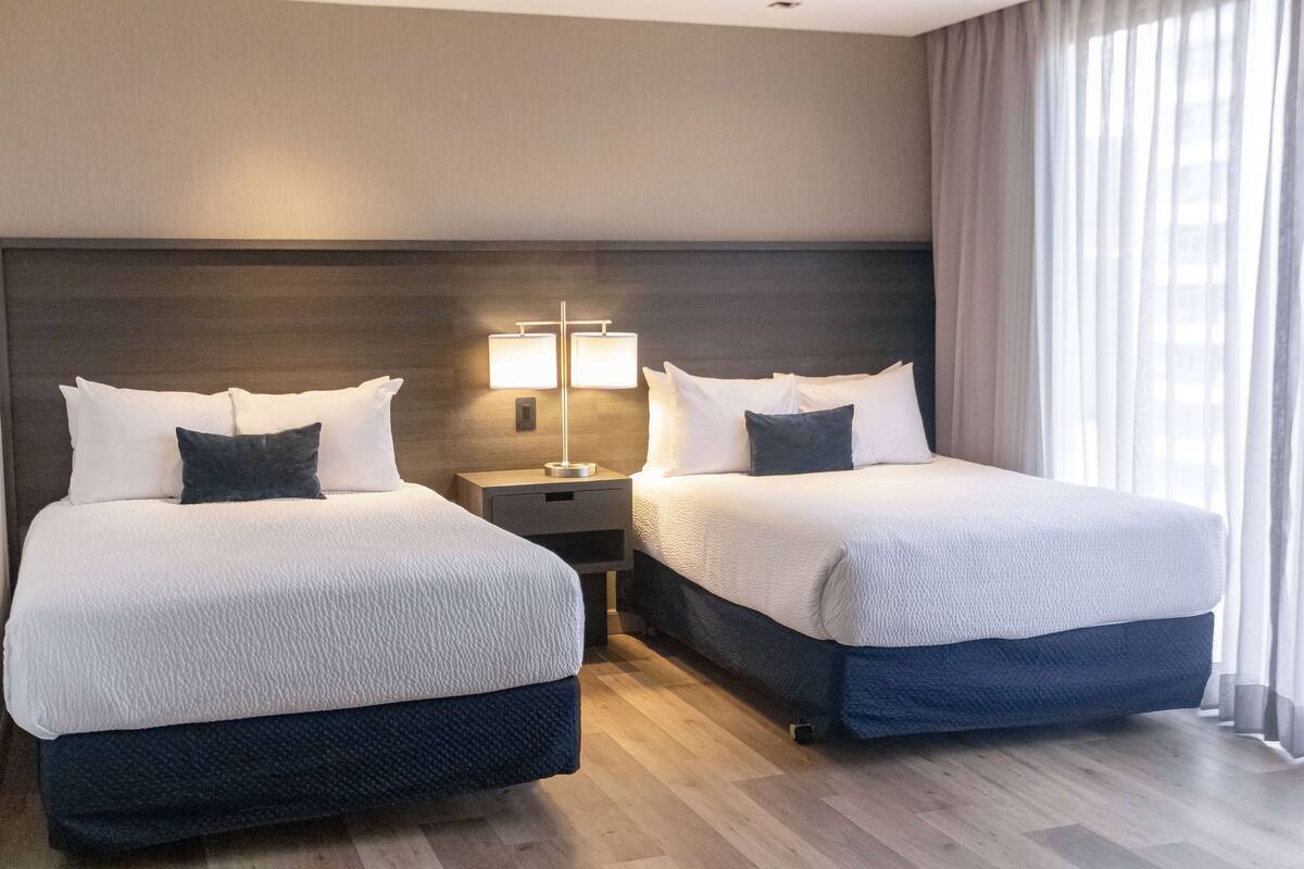 Ein Hotelzimmer mit zwei Betten, einem Nachttisch mit Lampe und einem großen Fenster mit Vorhängen.