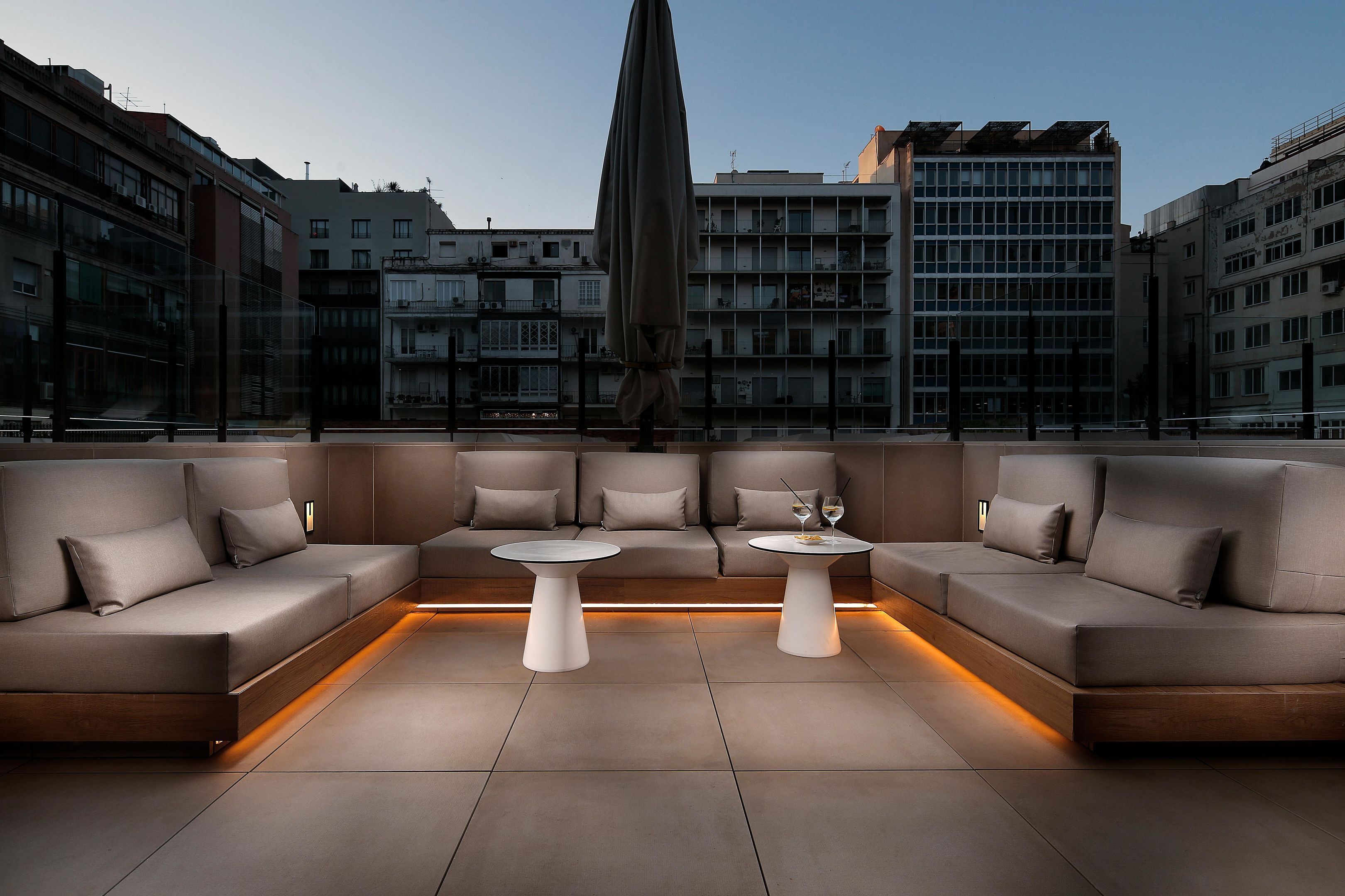 A modern outdoor seating area with a sofa and two tables, a large umbrella, and a cityscape view in the background.