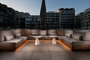 A modern outdoor seating area with a sofa and two tables, a large umbrella, and a cityscape view in the background. - HCC St. Moritz (Barcelona)