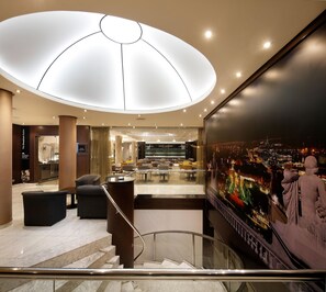 Un hall d’hôtel moderne, doté d’un grand lustre au plafond, de coins salon et offrant une vue sur le paysage urbain.