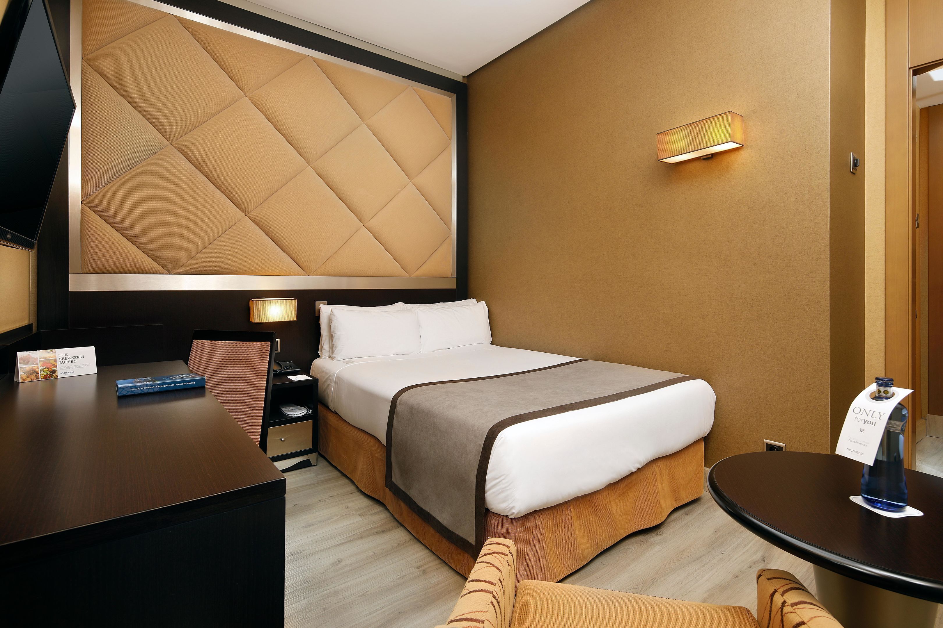 Habitación de hotel con una cama grande, un escritorio, una silla, una mesita de noche y un televisor instalado en la pared.