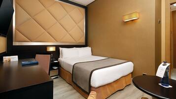 Habitación de hotel con una cama grande, un escritorio, una silla, una mesita de noche y un televisor instalado en la pared.
