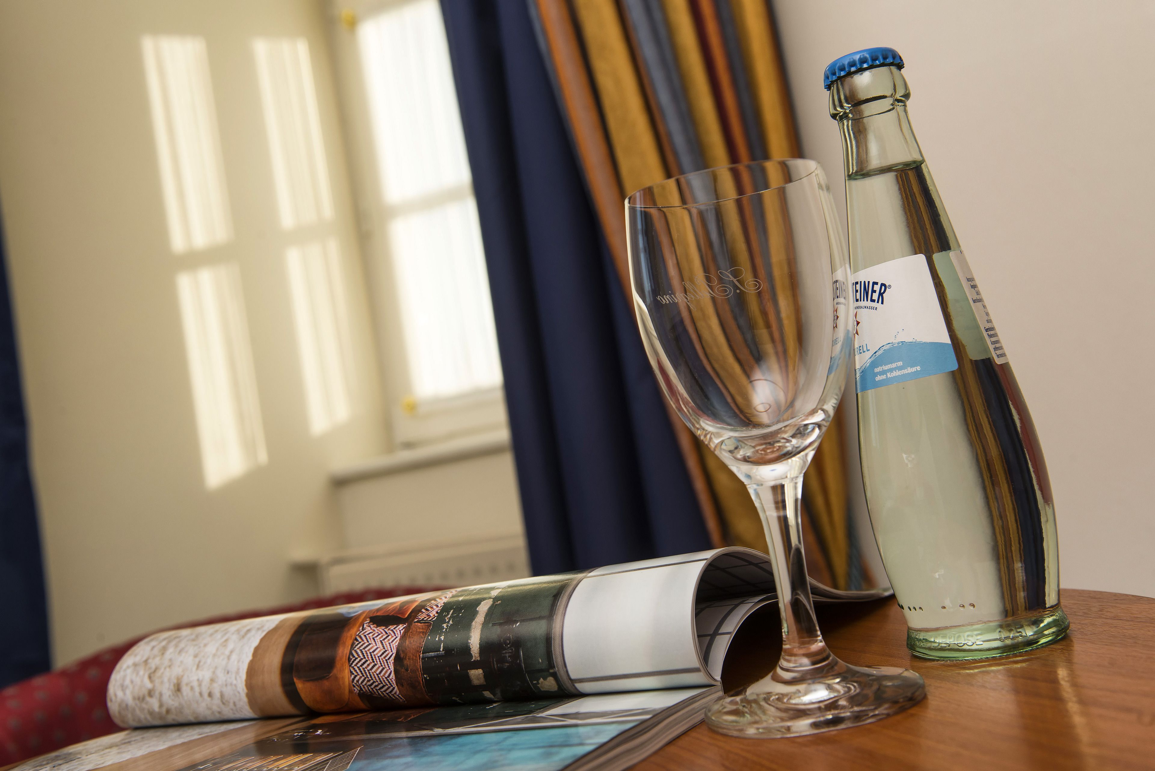 A clear glass bottle and a wine glass on a wooden table with a magazine.