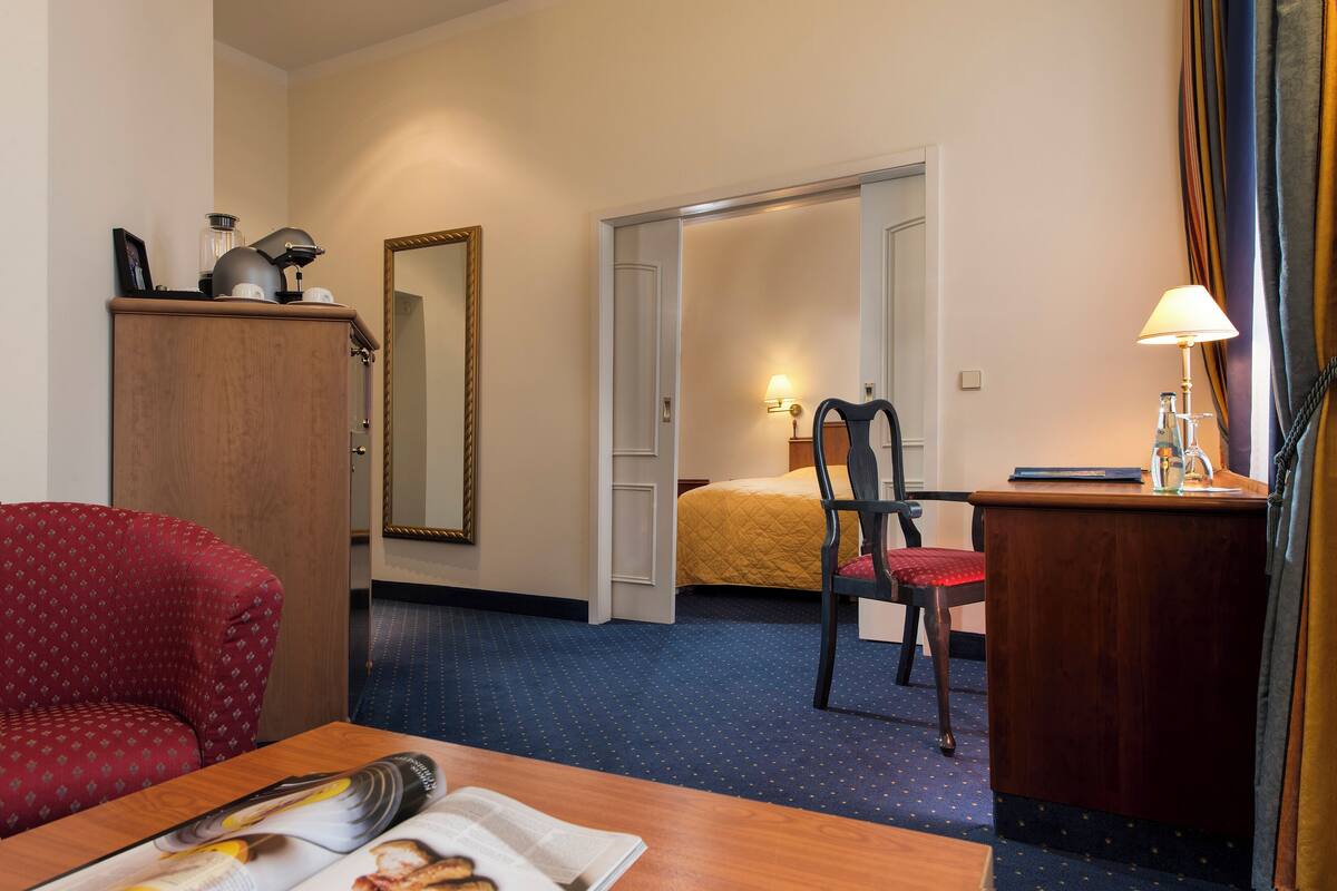 Una habitación de hotel con cama, escritorio, silla, espejo y un sillón rojo.