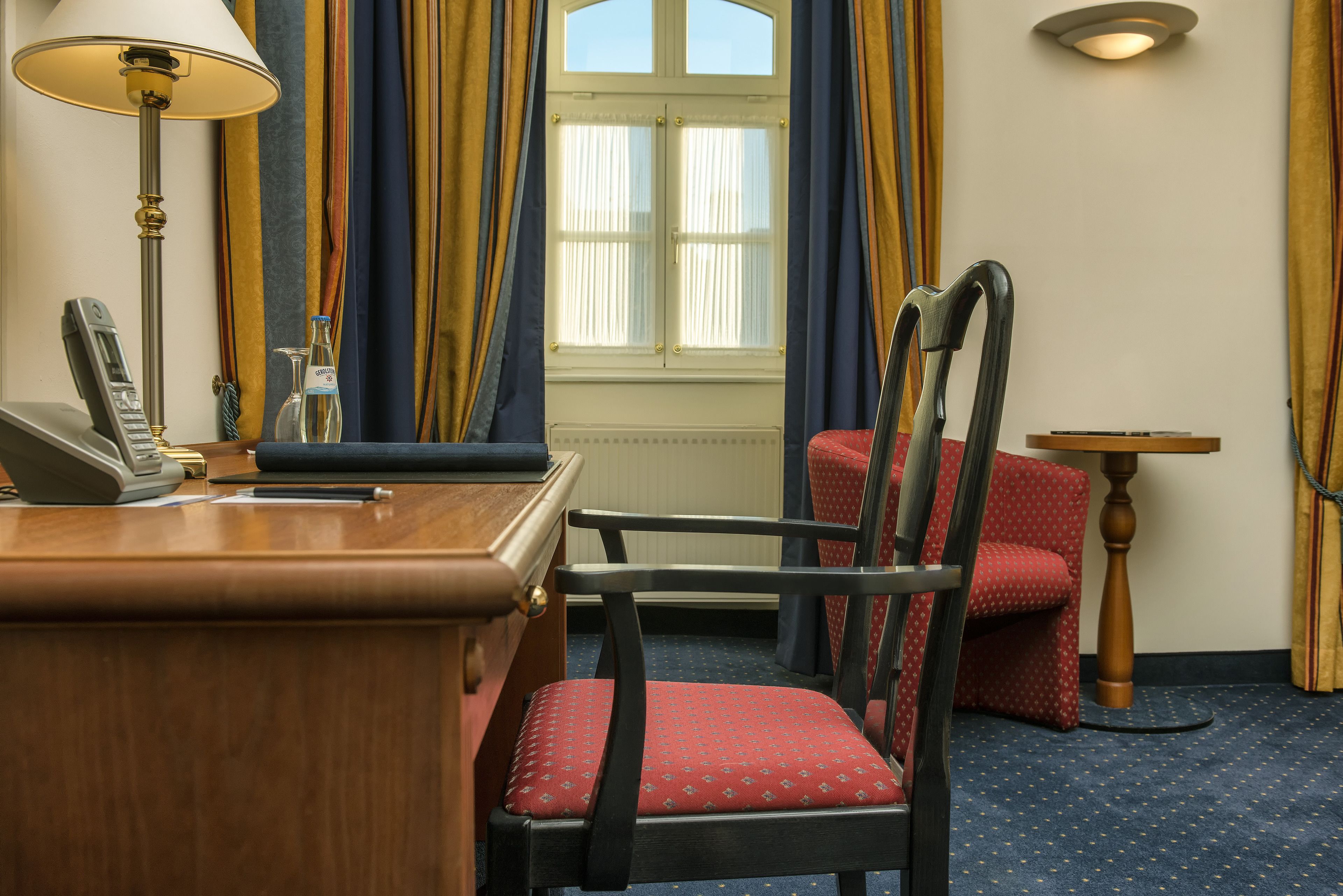 Une chambre d’hôtel avec un bureau en bois, un téléphone, une chaise avec un coussin rouge, une petite table et une fenêtre avec des rideaux.