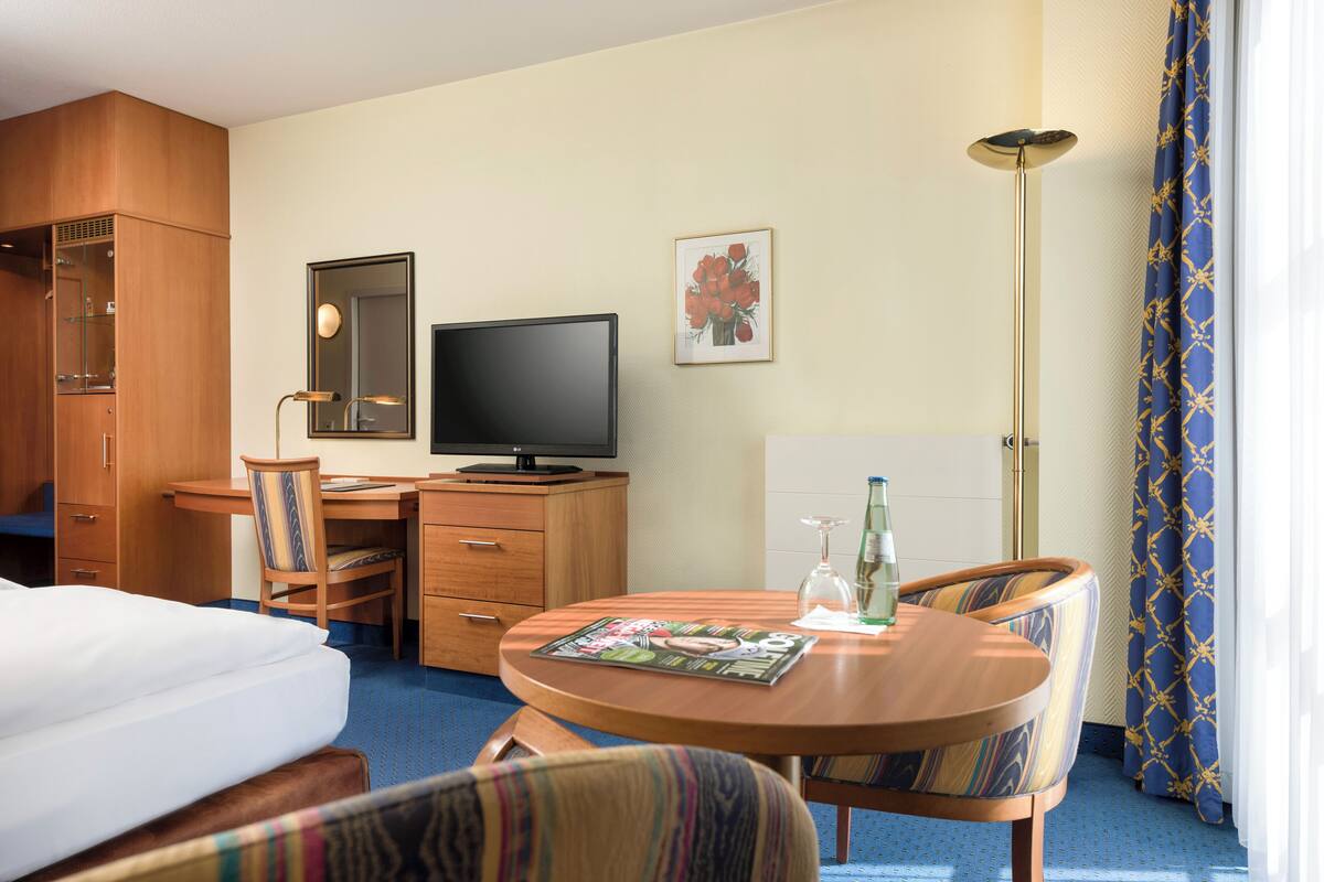 Habitación de hotel con cama, escritorio con computadora, silla, mesa con revista, lámpara y ventana con cortinas.