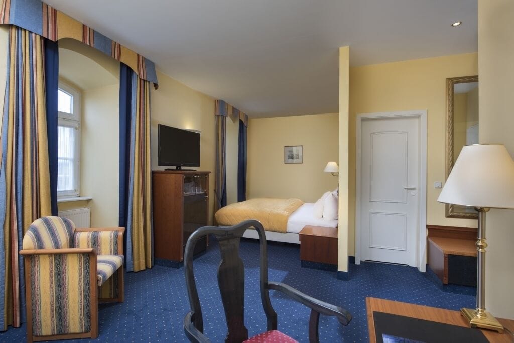 A hotel room with a bed, two armchairs, a desk with a lamp, a television, and a door.