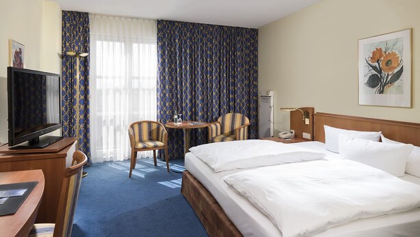 A hotel room with a bed, two chairs, a desk with a TV, a lamp, and a painting on the wall. - Radisson Blu Hotel, Halle-Merseburg (Merseburg)
