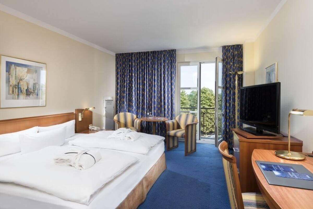 Habitación de hotel con dos camas, un escritorio con televisor, un balcón con cortinas y un cuadro en la pared.