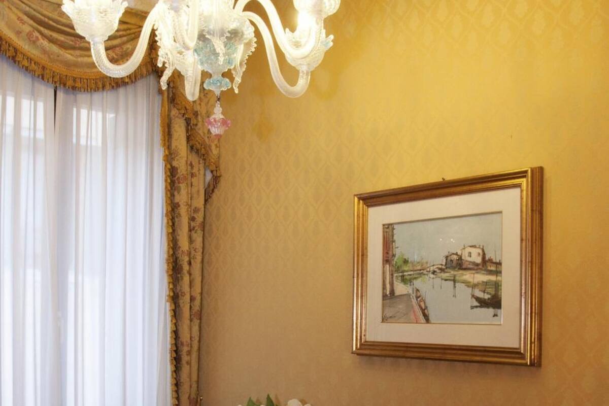 Une chambre d’h ôtel avec un lit, un bureau en bois, une chaise, un lustre, un tableau encadré et un vase avec des fleurs.