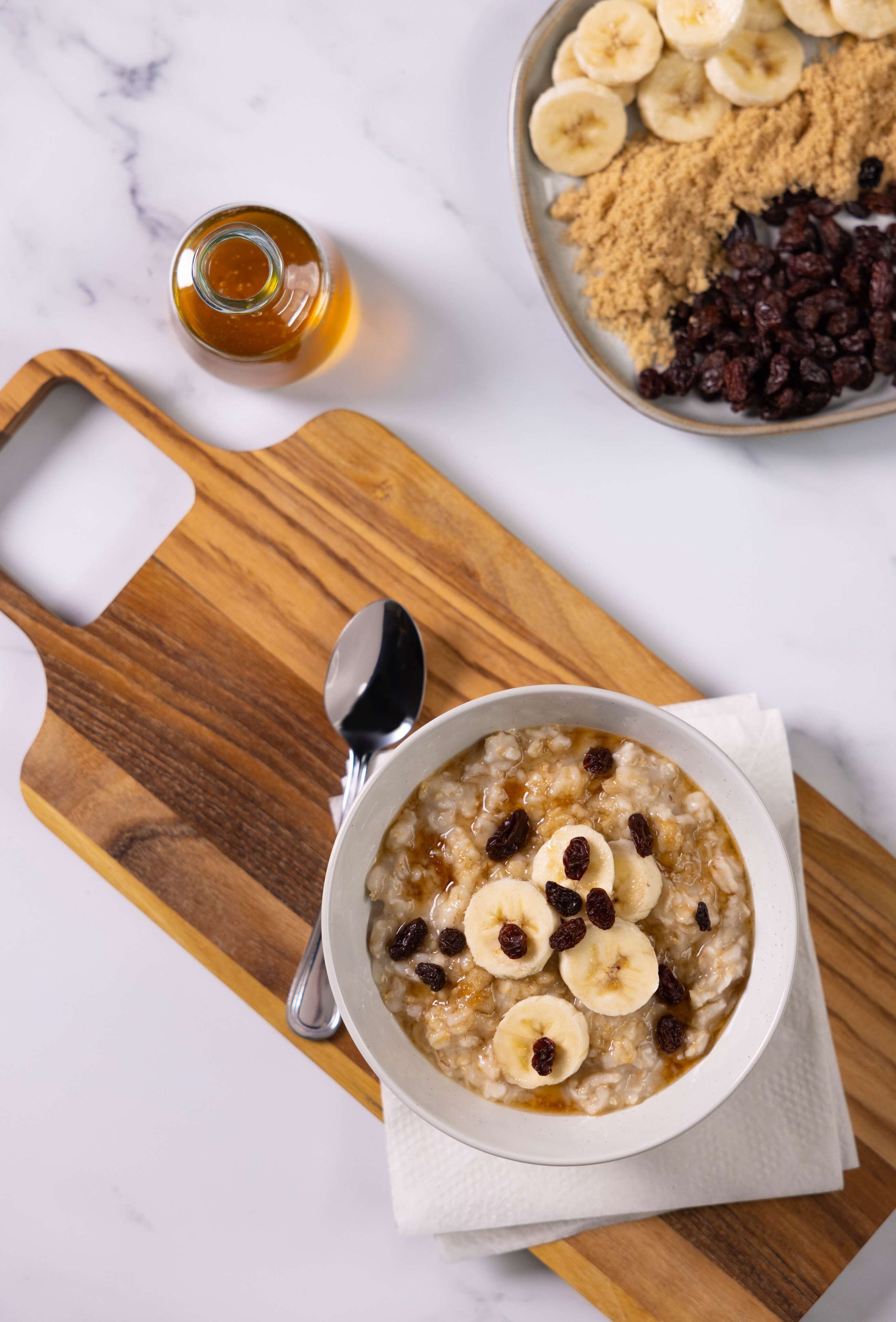 A bowl of oatmeal with banana slices and raisins on a wooden board, with a spoon and a bottle of syrup nearby.