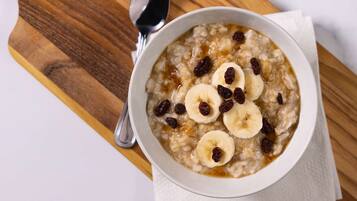 A bowl of oatmeal with banana slices and raisins on a wooden board, with a spoon and a bottle of syrup nearby.