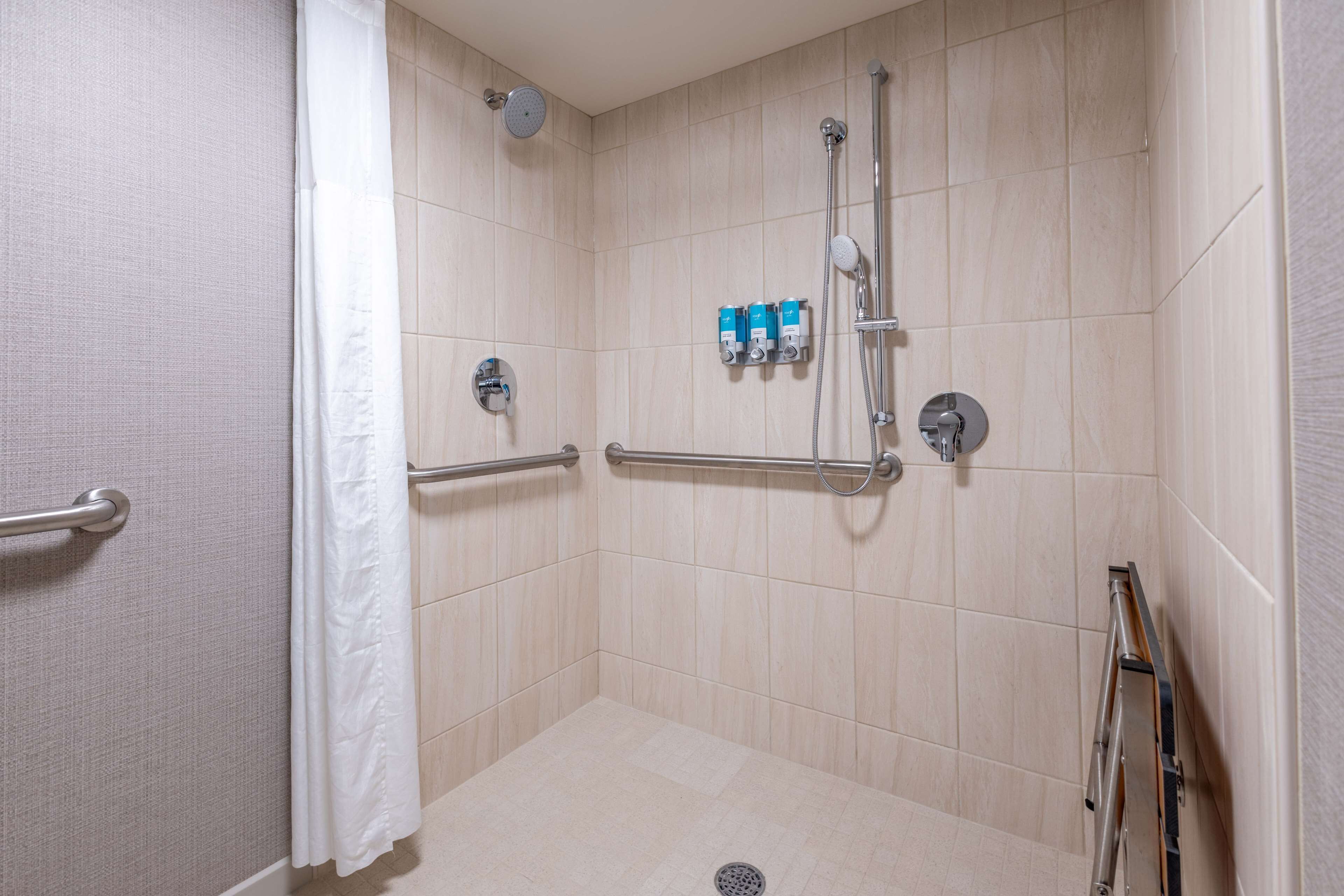 A walk-in shower with beige tiles, a grab bar, a handheld showerhead, and a handheld showerhead with a hose.
