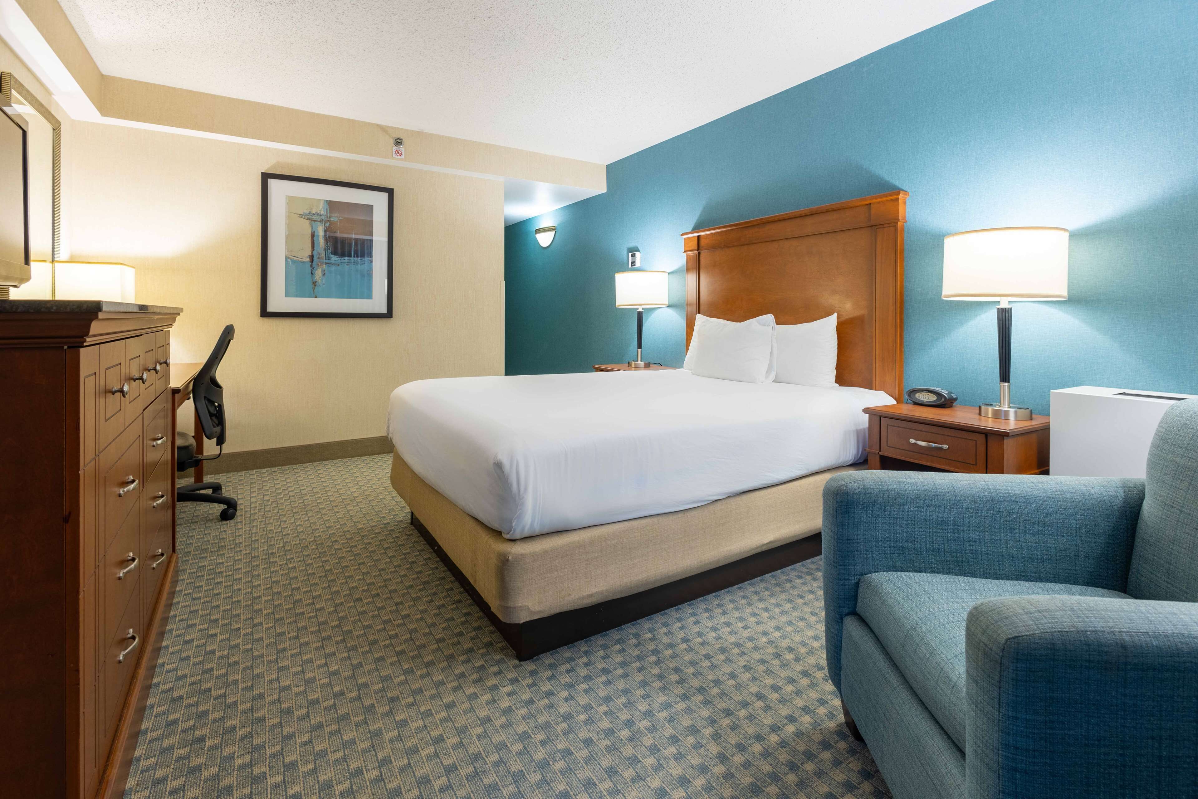 A hotel room with a large bed, a wooden headboard, a desk with a lamp, a chair, and a dresser.