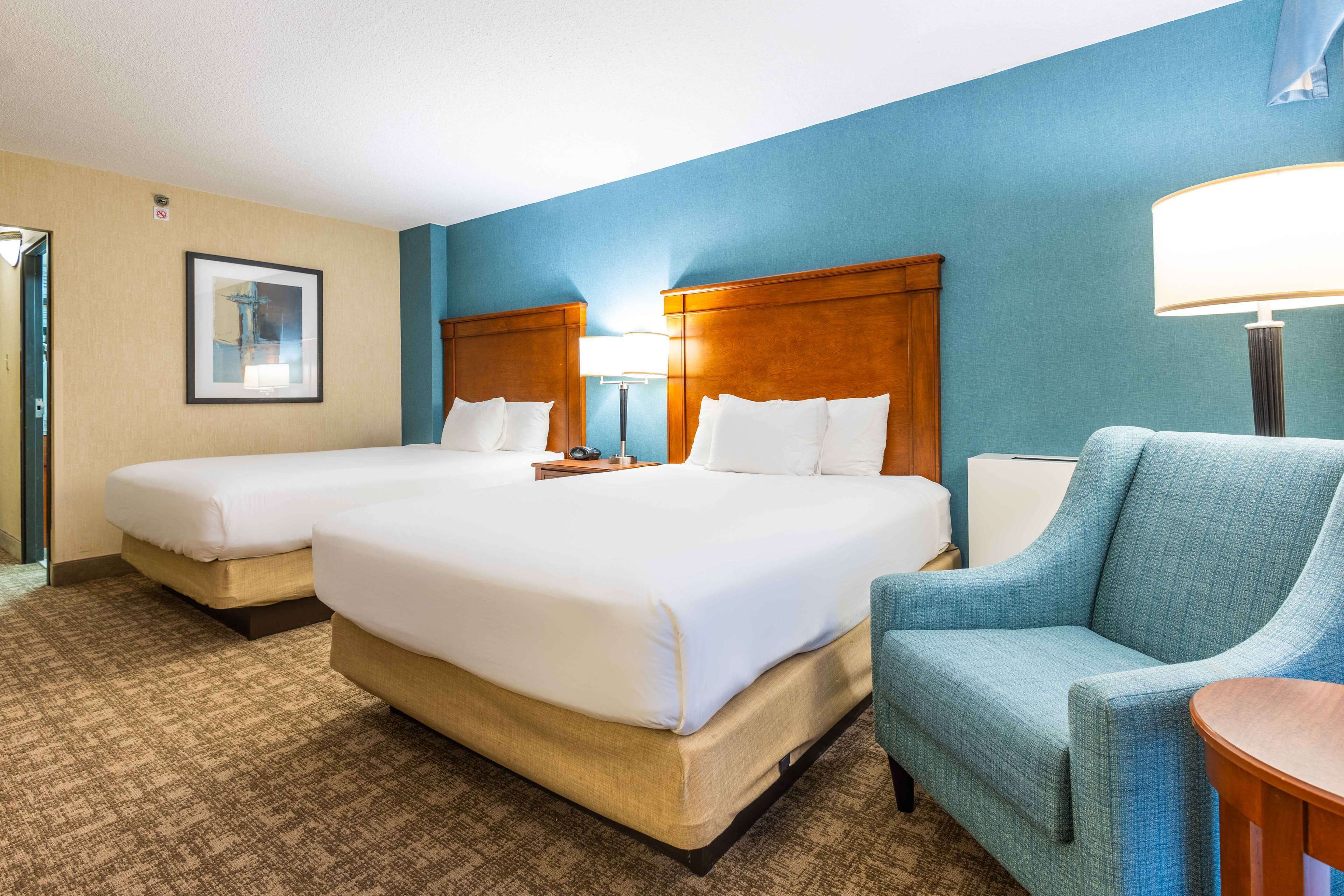 A hotel room with two beds, a blue wall, a wooden headboard, a blue armchair, and a lamp.
